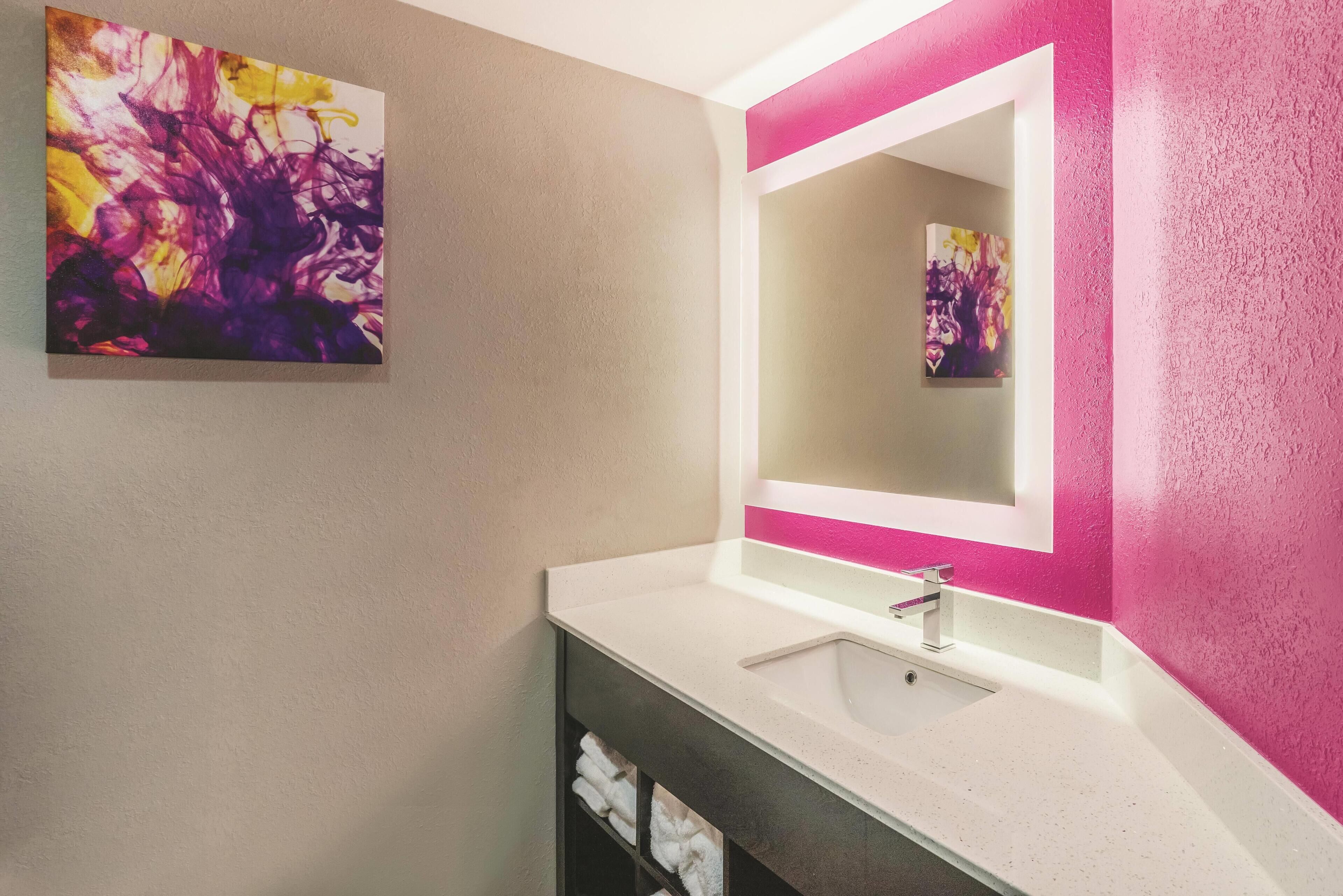 Un bagno con un lavandino bianco, uno specchio con cornice rosa e un quadro astratto appeso alla parete.