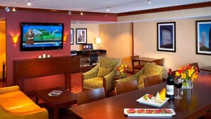 A hotel room with a large bed, a desk with a computer, two patterned armchairs, a dining table with a bottle of wine and flowers, and a TV displaying a weather forecast.