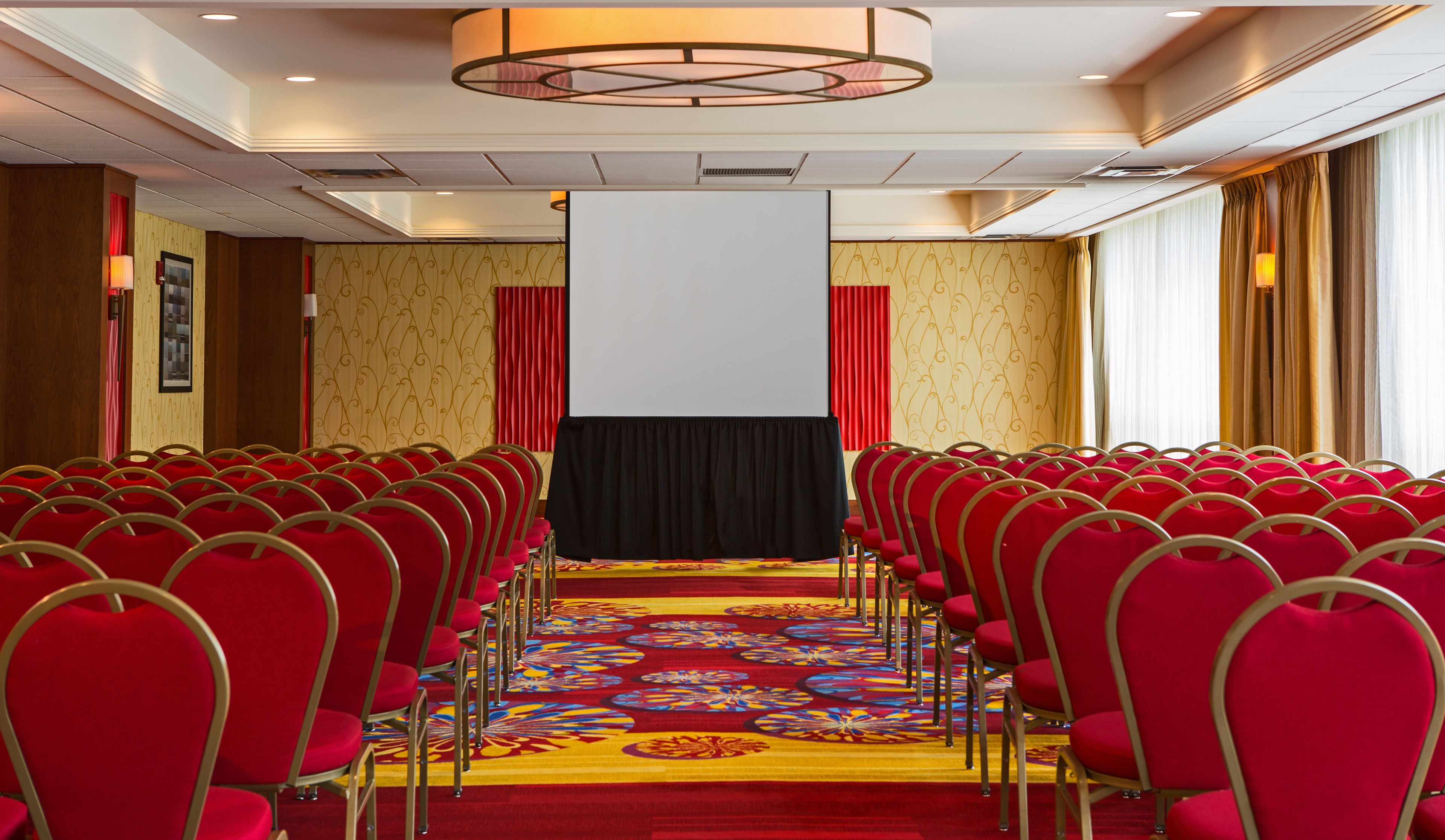 Ein Konferenzraum mit Reihen roter Stühle, einem großen Teppich mit Blumenmuster und einer weißen Leinwand im Vordergrund.