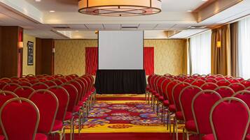Un salón de conferencias con filas de sillas rojas, una alfombra grande con motivos florales y una pantalla blanca al frente.