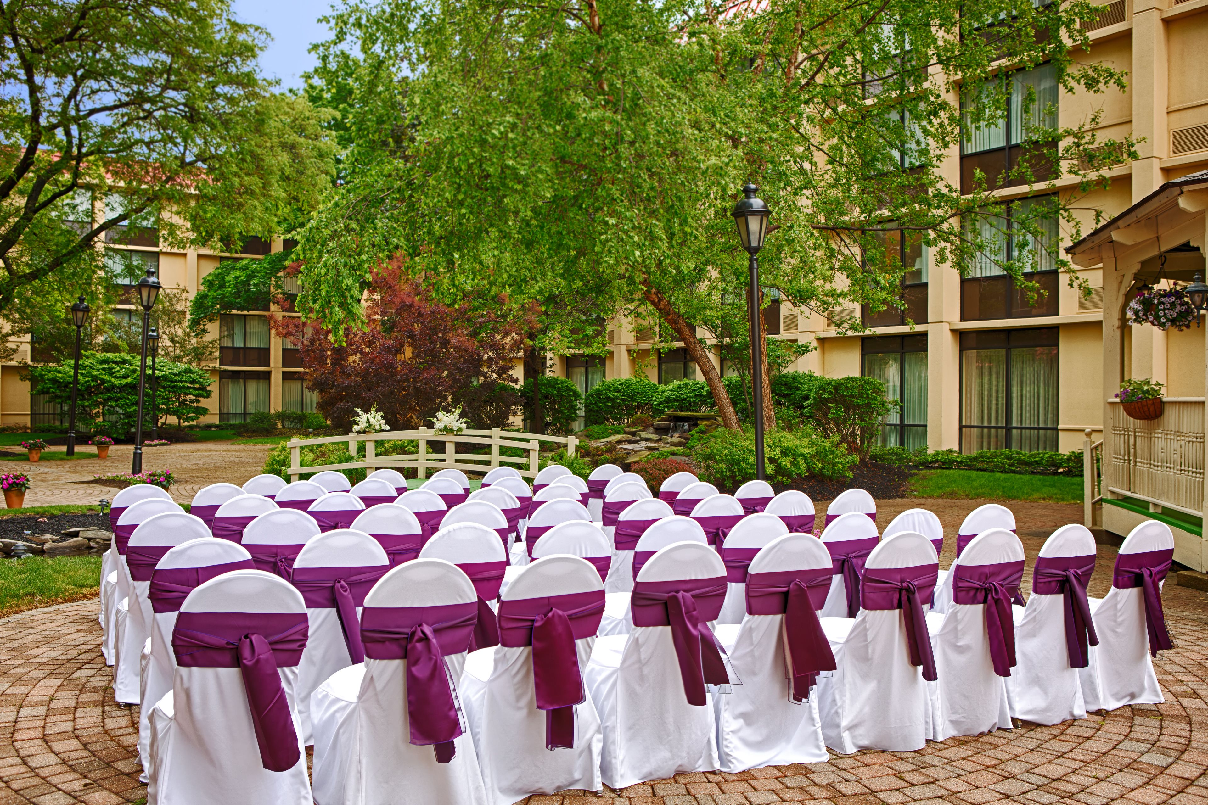 Rows of white chairs with purple sashes arranged outdoors, surrounded by greenery and a building with balconies.