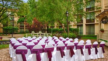 Rows of white chairs with purple sashes arranged outdoors, surrounded by greenery and a building with balconies.