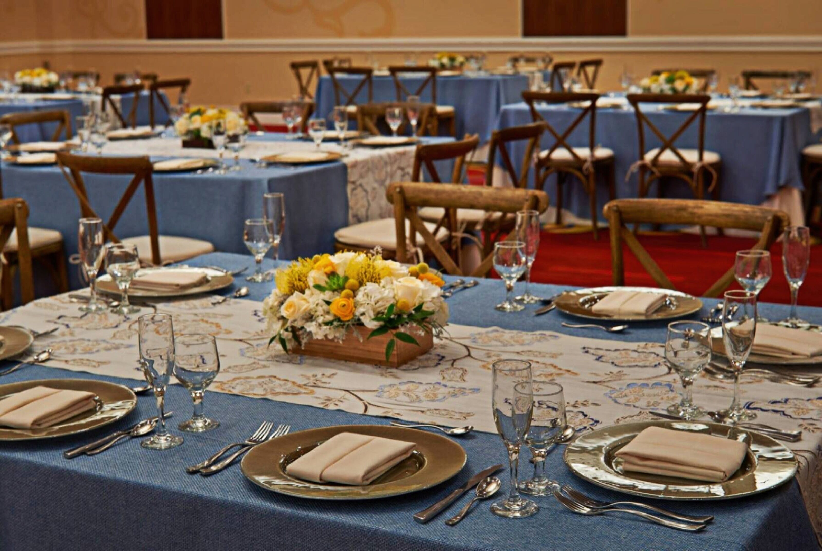 Un salón de banquetes con mesas redondas dispuestas para recibir comensales, cada una con mantel azul, servilletas blancas y centros de mesa florales.