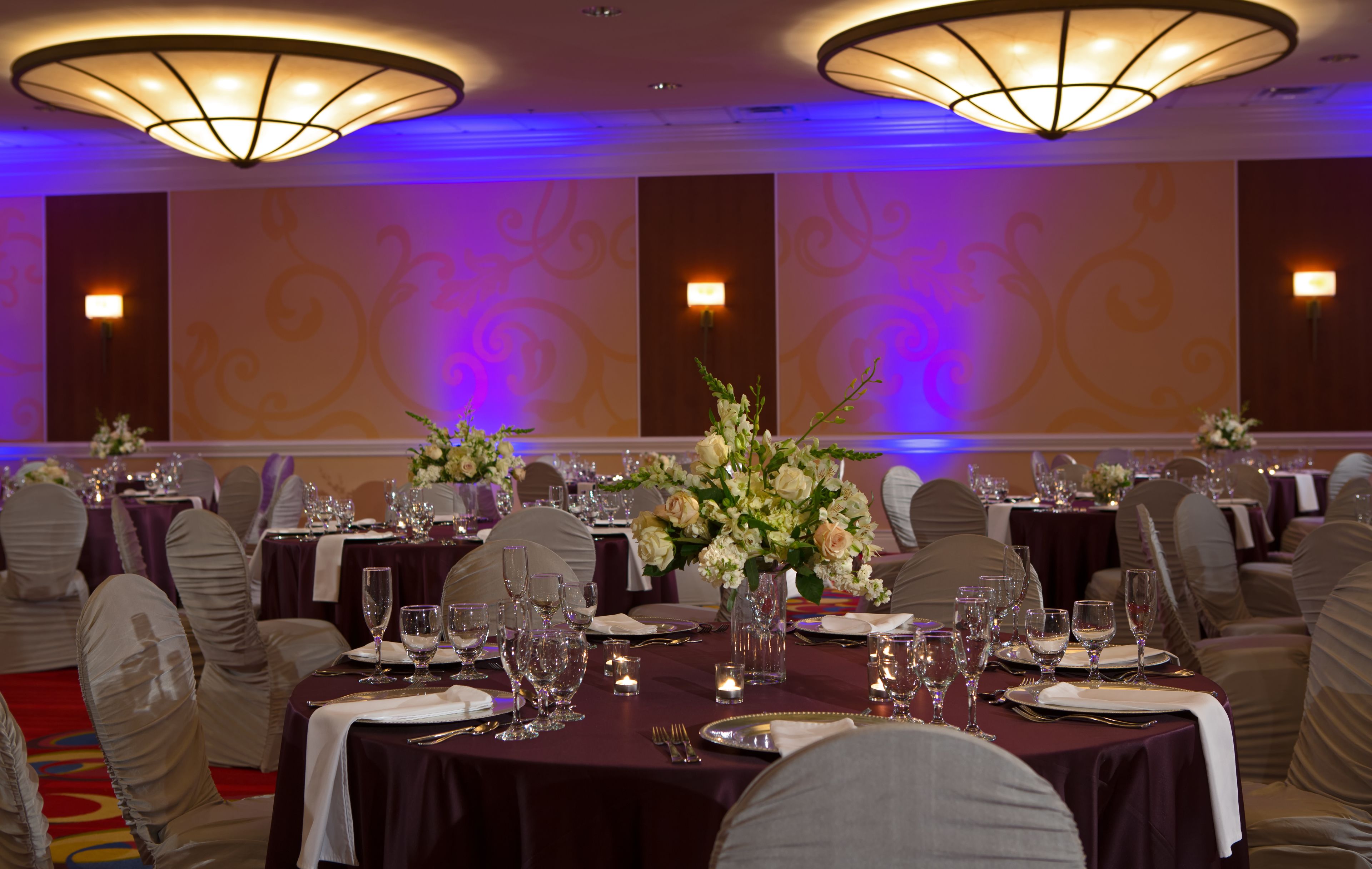 A banquet hall with round tables set for dining, each with a vase of flowers, and overhead lighting fixtures.