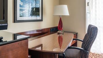 A hotel room with a wooden desk, a red lamp, a framed picture on the wall, and a patterned chair.