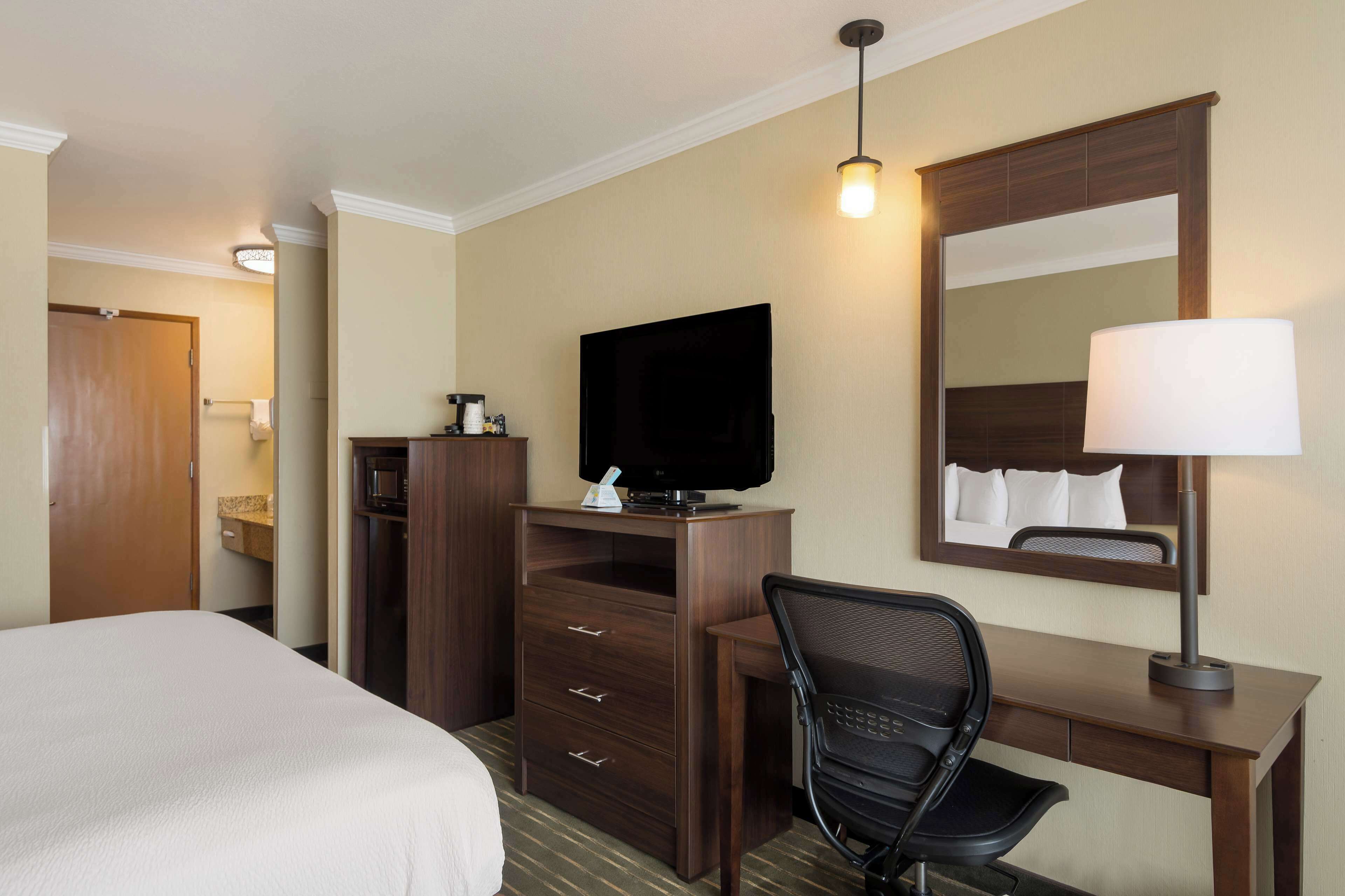 A hotel room with a bed, a desk with a chair, a television, a mirror, and a lamp.