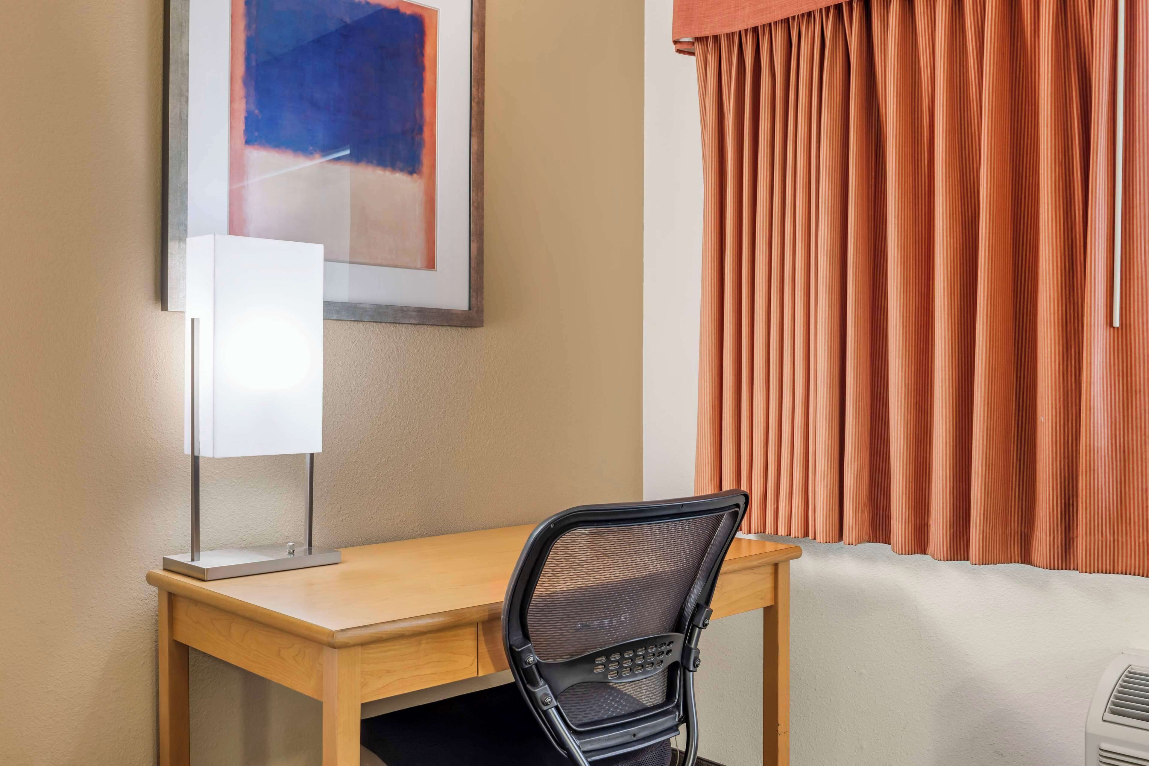 Une chambre d’hôtel avec un bureau en bois, une chaise noire à dossier en tissu grillagé, un tableau abstrait encadré et des rideaux orange.
