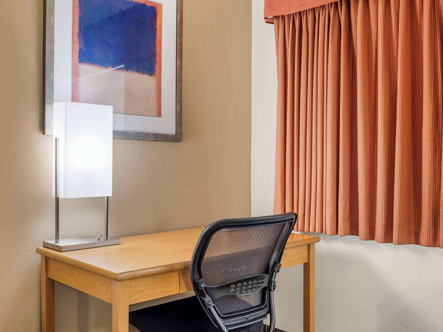 A hotel room with a wooden desk, a black mesh chair, a framed abstract painting, and orange curtains.