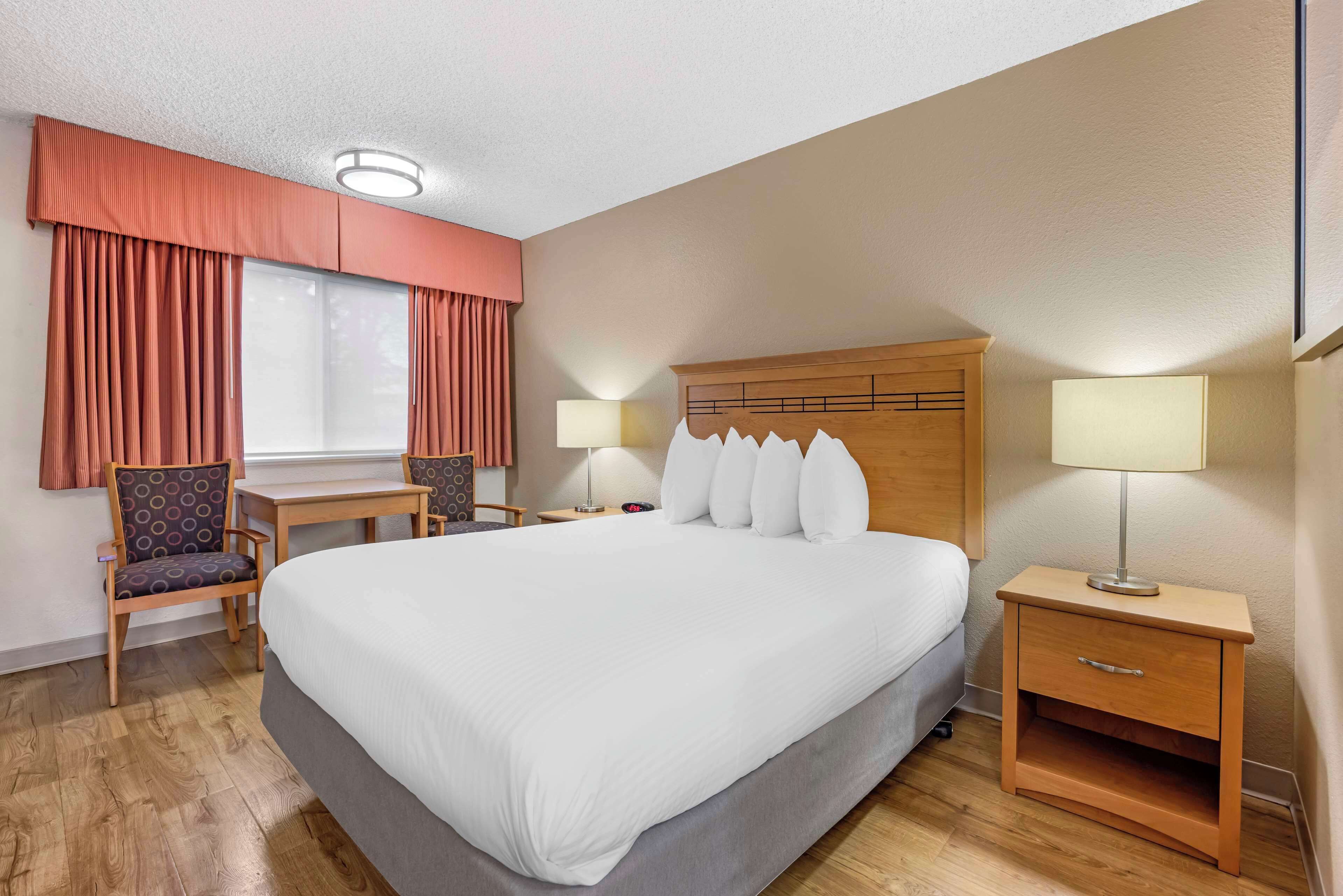A hotel room with a large bed, two bedside tables with lamps, a desk with a chair, and a window with curtains.