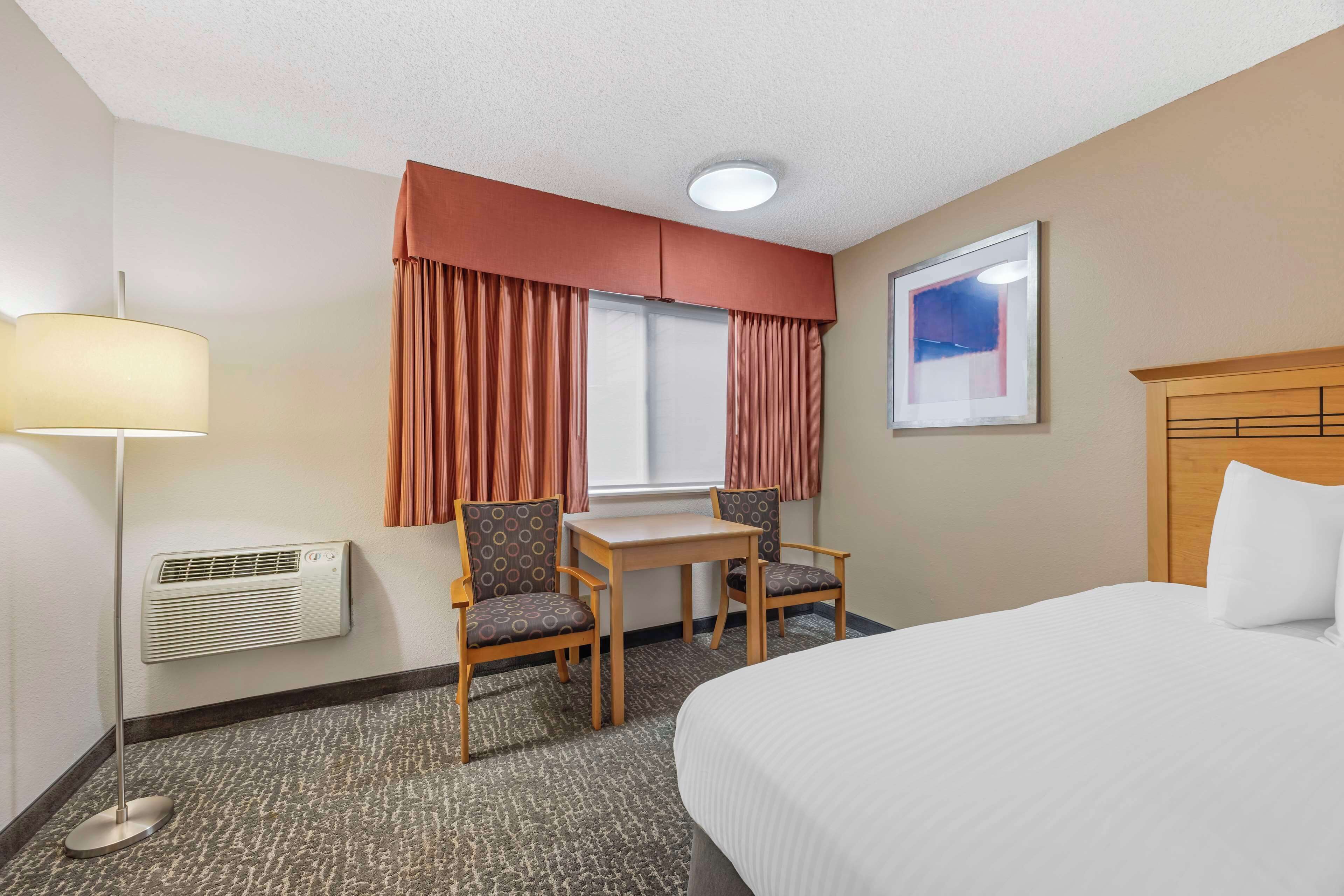 A hotel room with a bed, a desk with two chairs, a window with curtains, a wall-mounted air conditioner, and a framed picture on the wall.