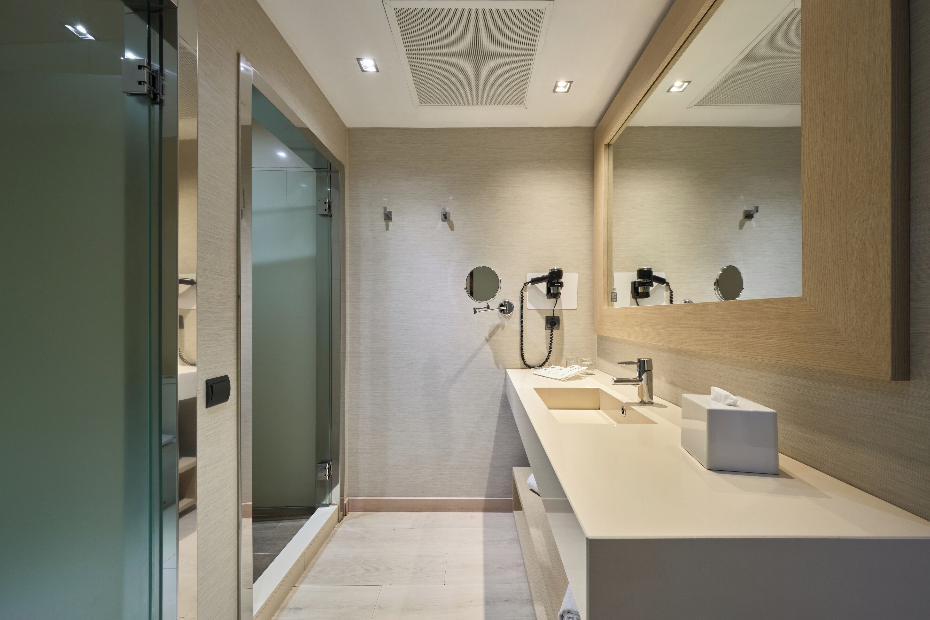 A modern bathroom with a glass shower enclosure, a large mirror, and a white vanity with a towel dispenser.