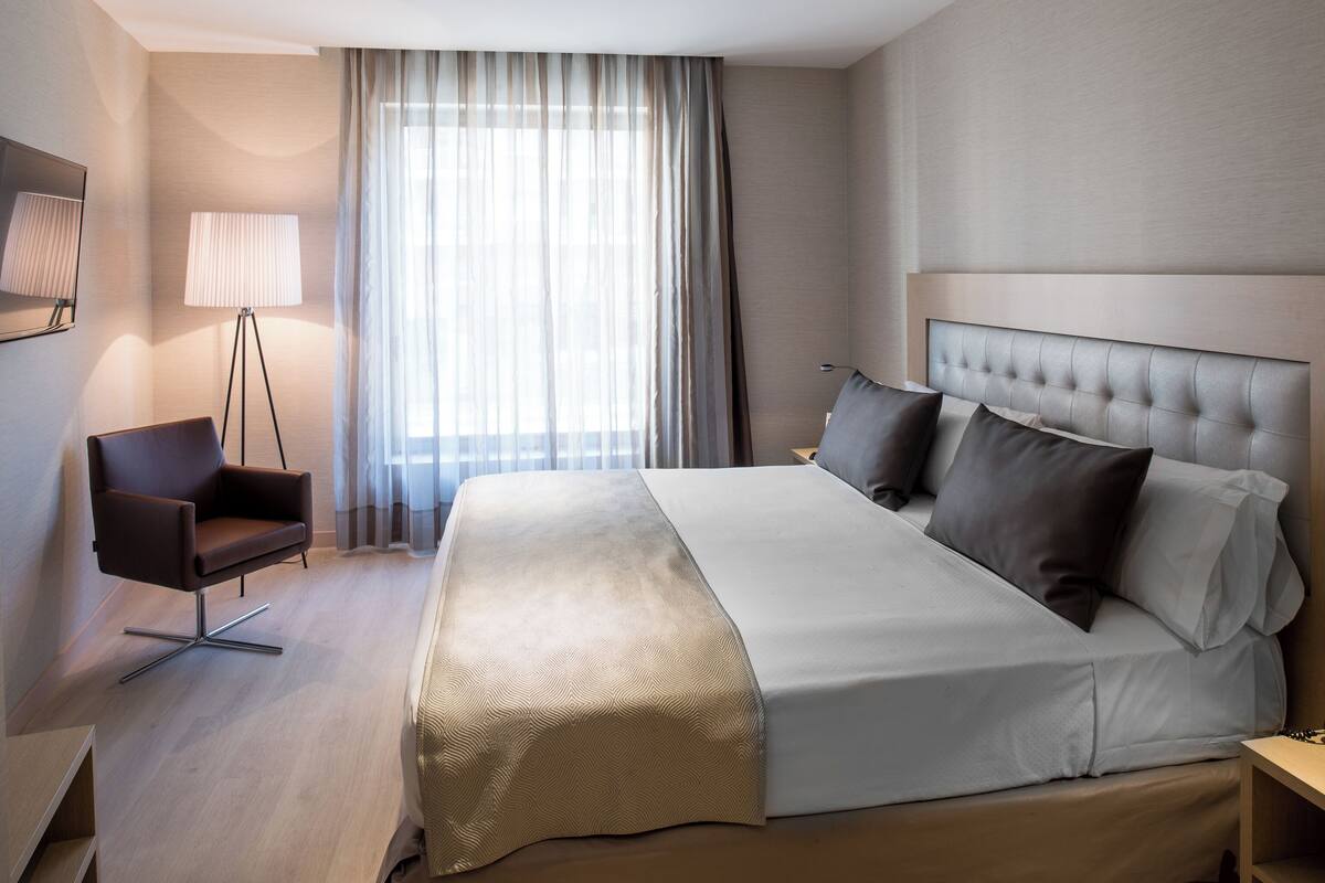 Quarto de hotel moderno com uma cama grande, uma cadeira, um abajur e vista para o exterior através de cortinas leves.