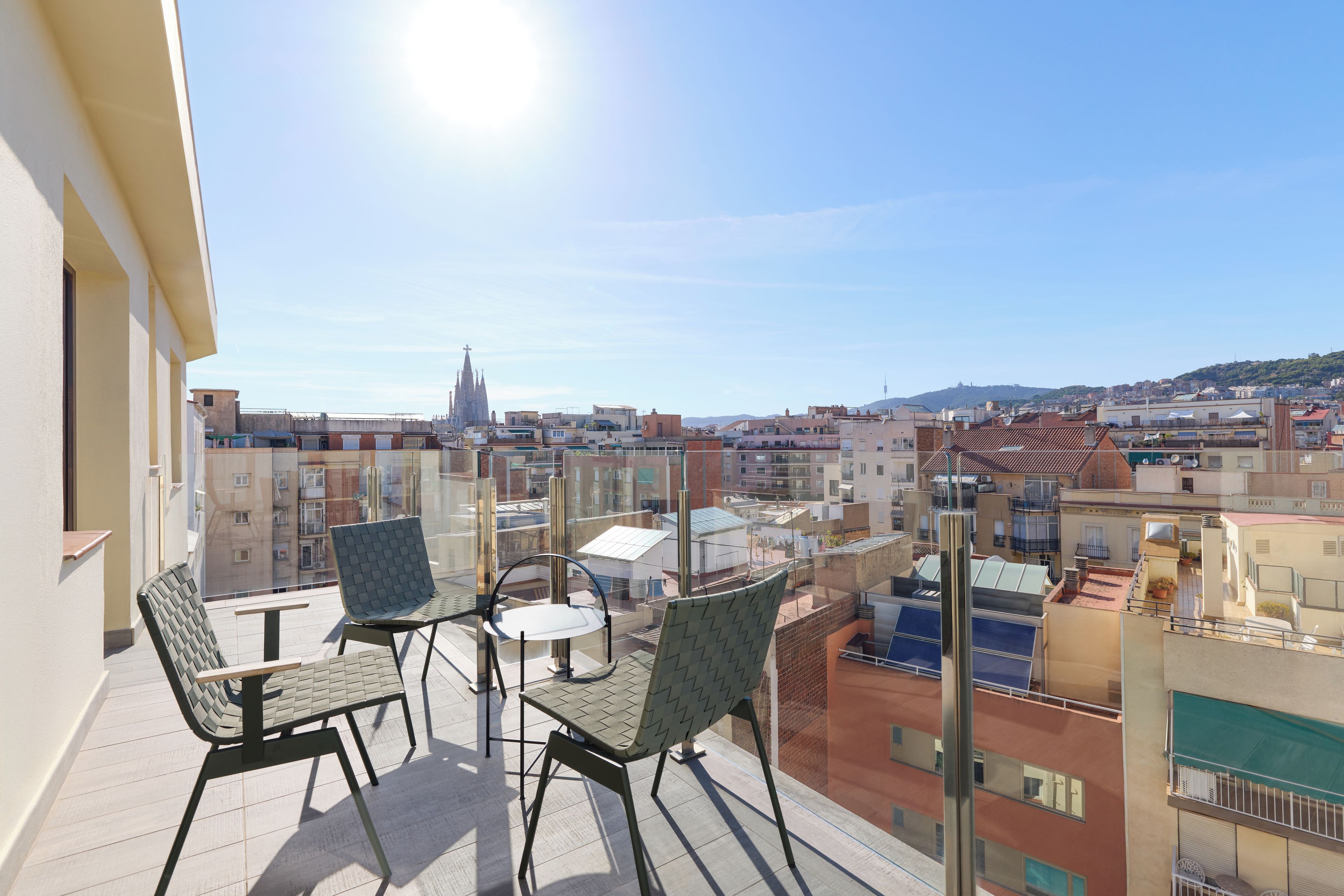 Un balcón con dos sillas y una mesa, con vistas a un paisaje urbano que incluye edificios y la aguja de una iglesia.