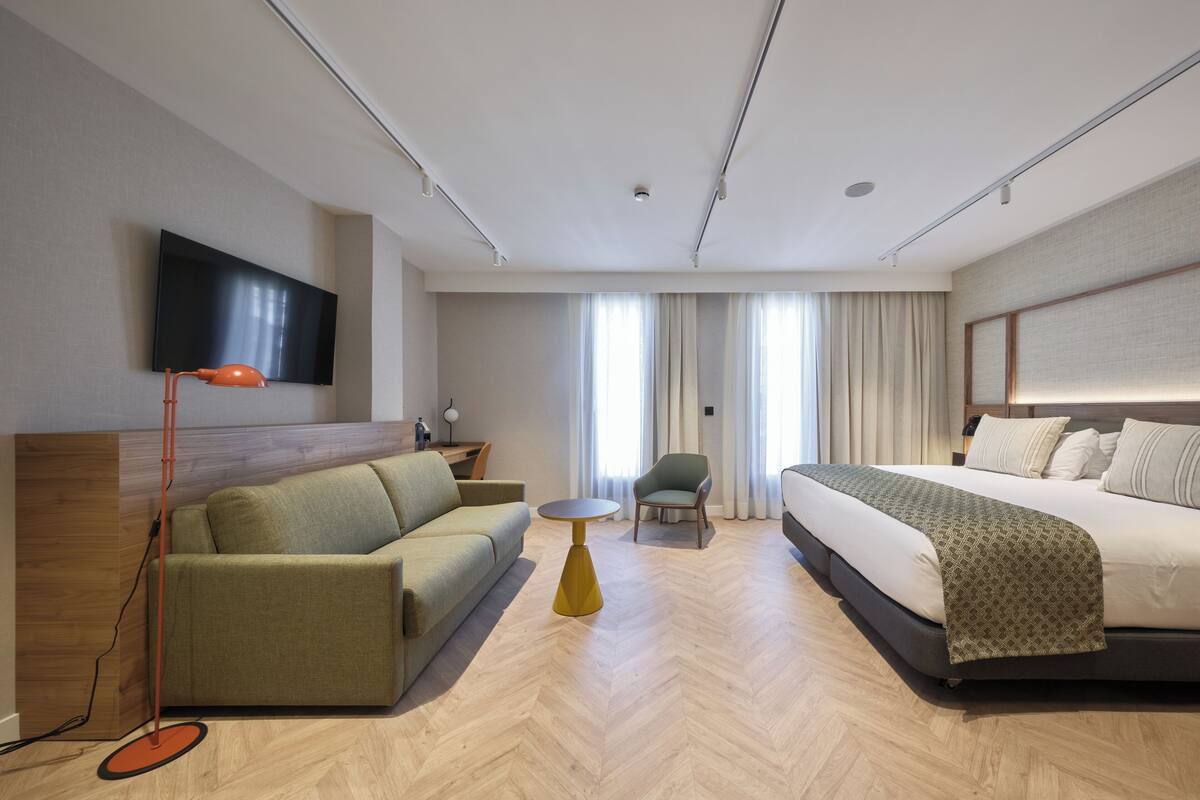 Quarto de hotel moderno com sofá, poltrona, cama, televisão e uma pequena mesa.