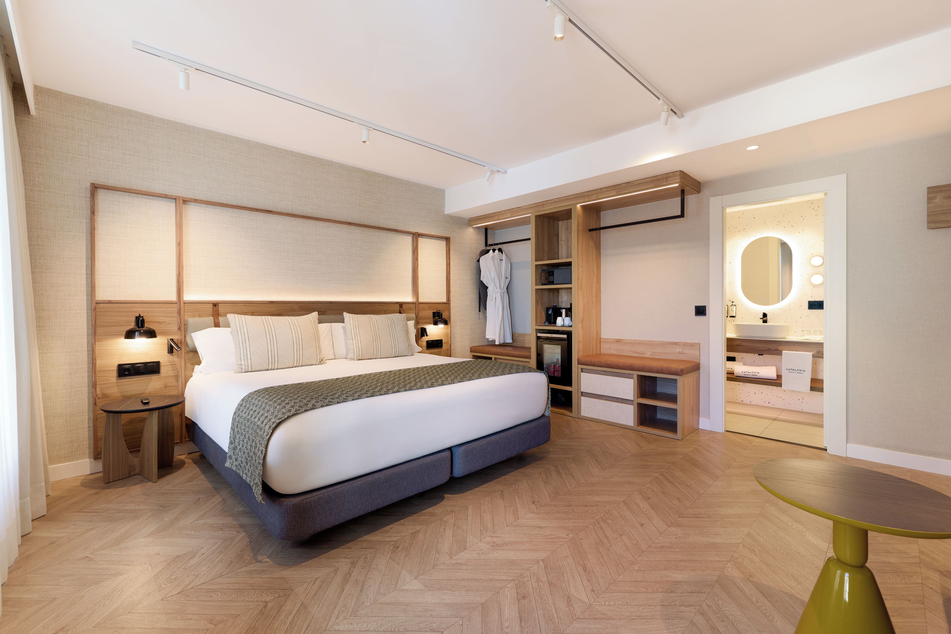 A modern hotel room with a large bed, bedside tables, a vanity with a mirror, and a seating area.