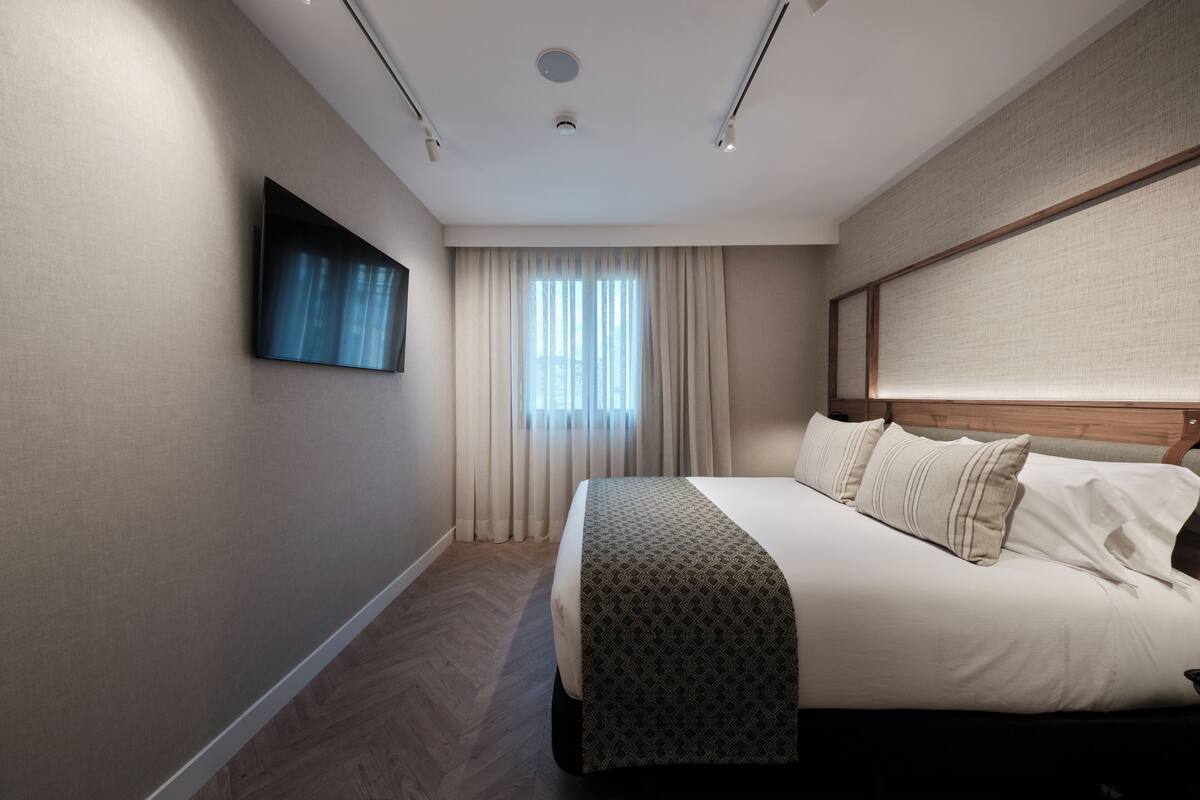 Quarto de hotel moderno com uma cama grande, uma TV de tela plana na parede e uma janela com cortinas translúcidas.