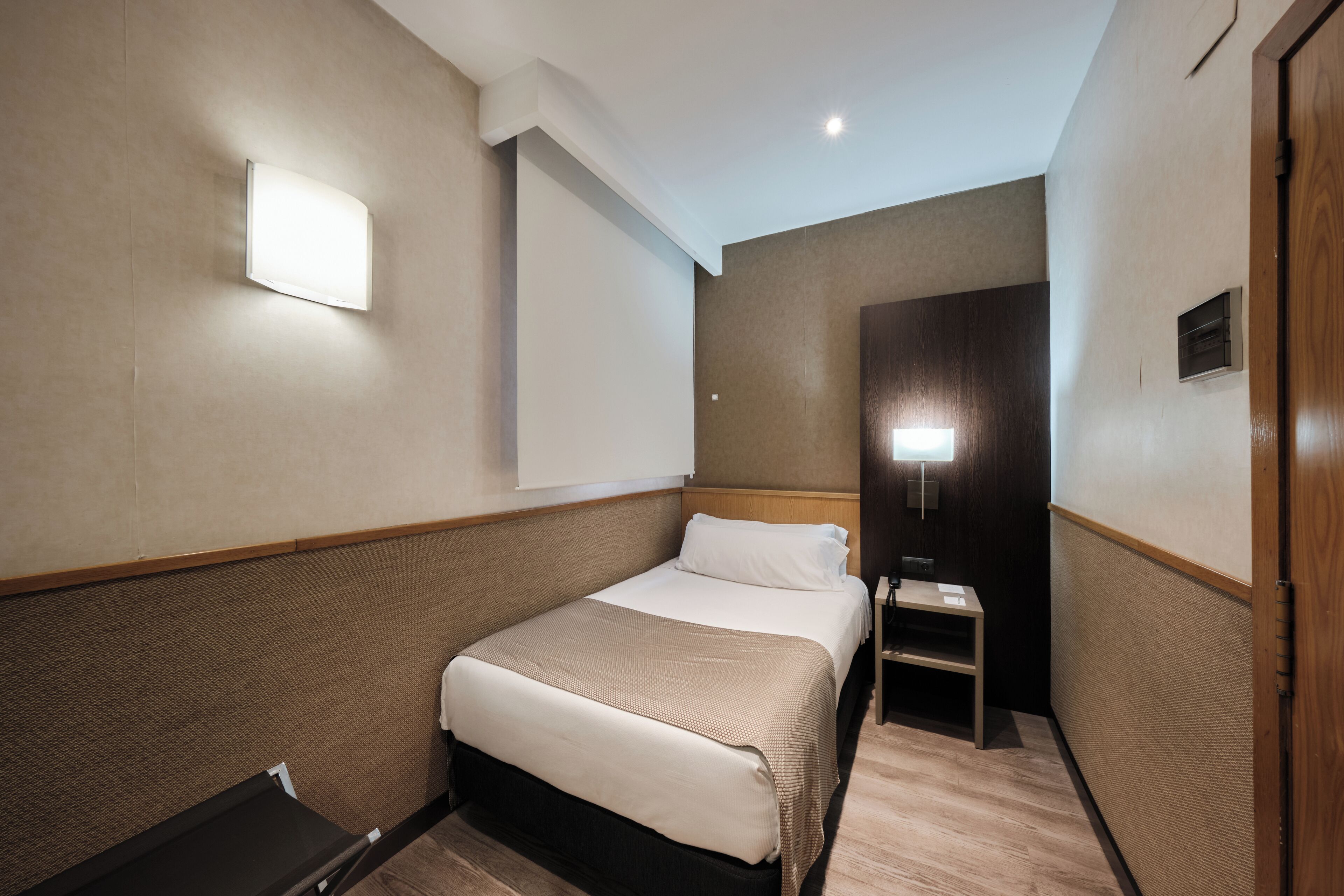 Une chambre d’hôtel avec un lit, une table de chevet, une lampe fixée au mur et une porte en bois.