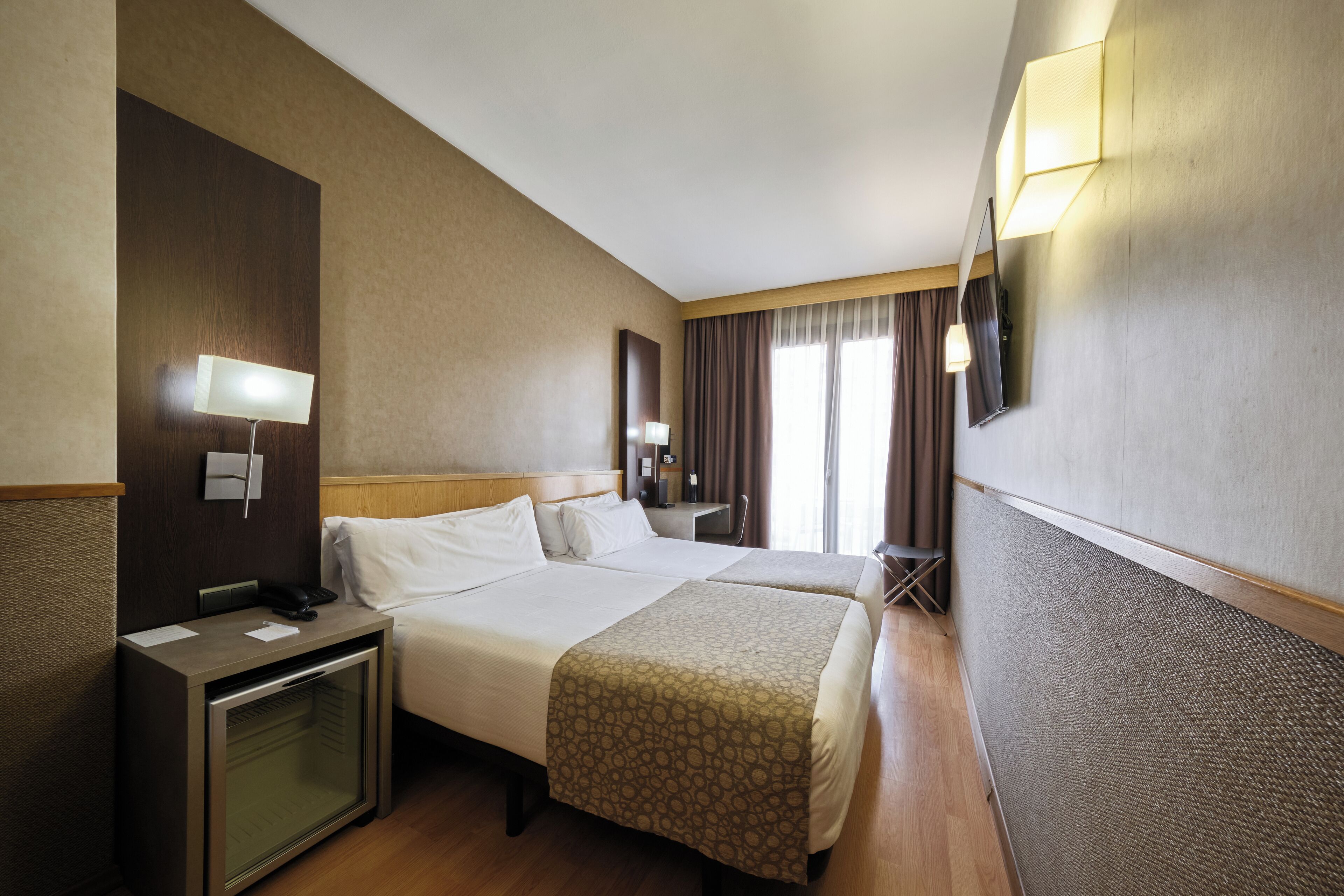 Habitación de hotel con dos camas, un escritorio con lámpara, un televisor montado en la pared y una ventana con cortinas.