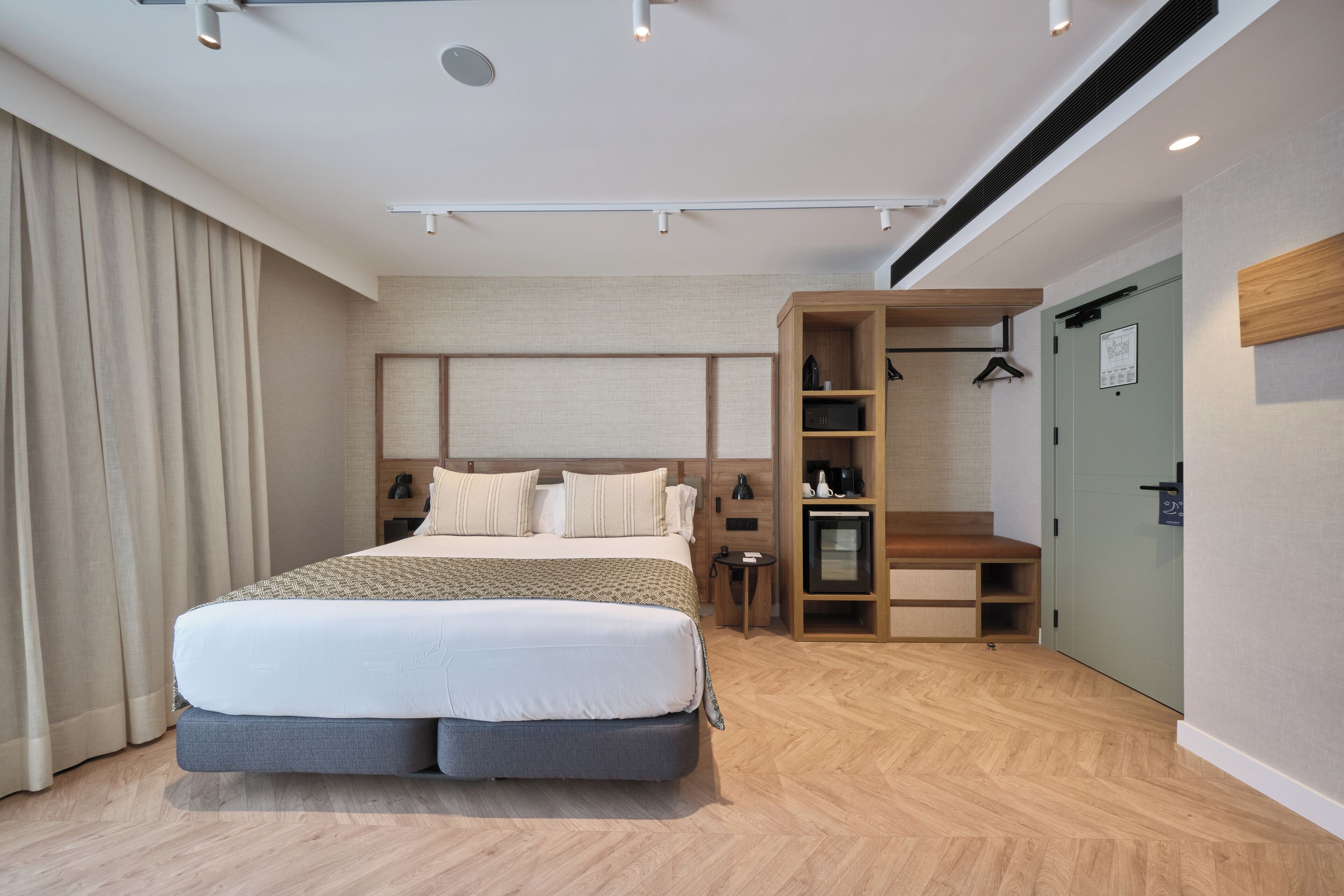 A modern hotel room with a large bed, a wooden bench, a desk, and a wardrobe.