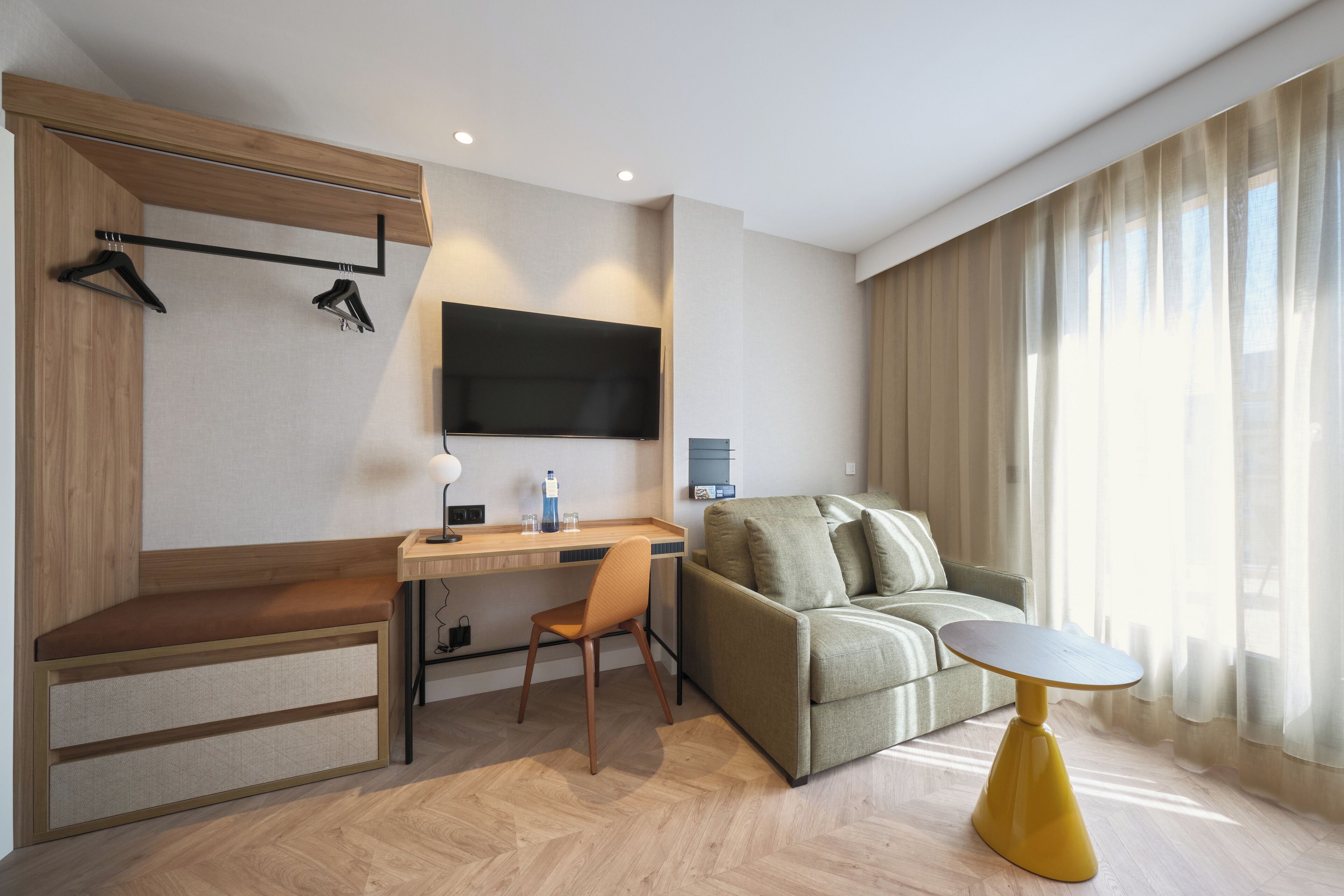 A modern hotel room with a wooden bench, a desk with a chair, a sofa, a small table, and a TV mounted on the wall.