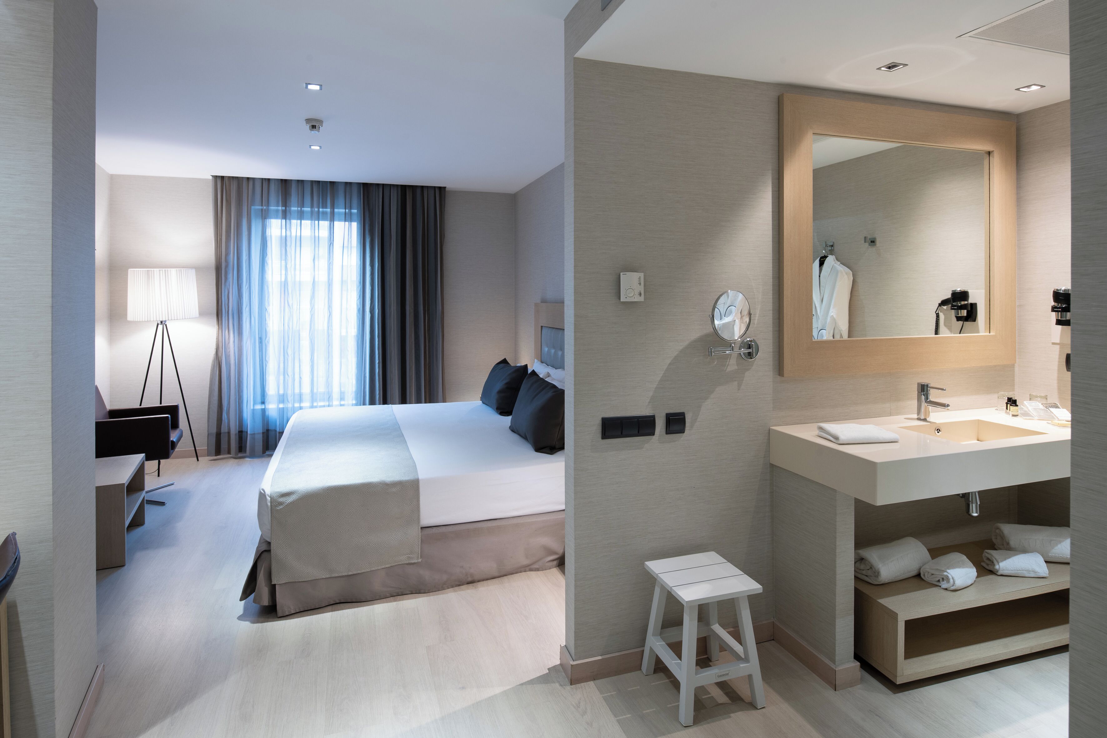 Una habitación de hotel moderna con una cama grande, un escritorio, una lámpara, un espejo y un baño con lavamanos y toallas.
