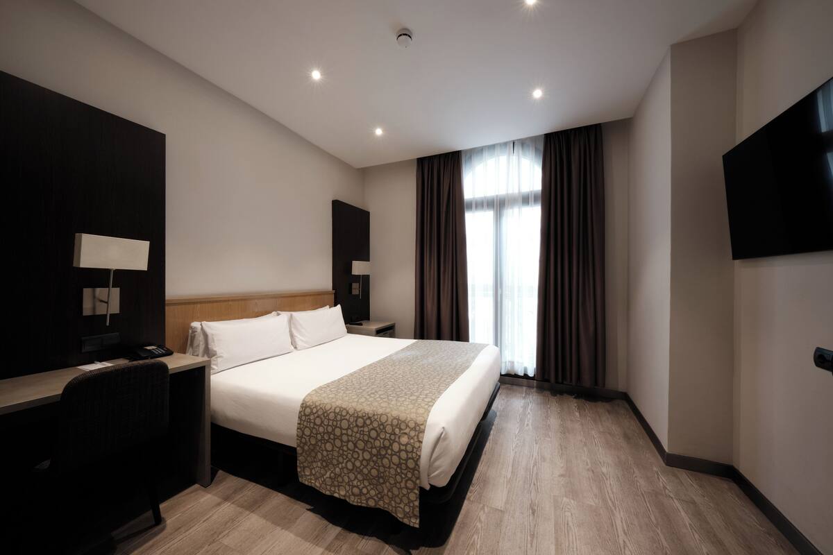 Quarto de hotel com uma cama grande, uma escrivaninha com abajur, televisão de tela plana e janela com cortinas.