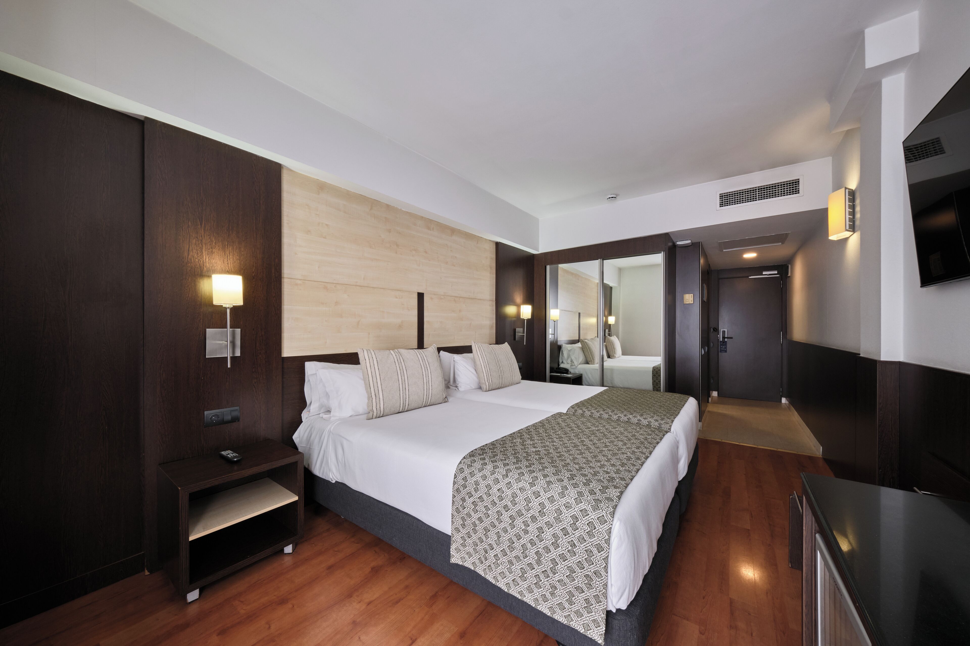 A modern hotel room with a large bed, bedside tables, a built-in TV, and a wooden floor.