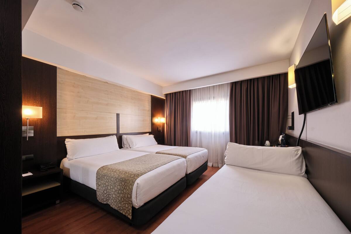 Quarto de hotel com duas camas, televisão, escrivaninha e uma janela grande com cortinas.