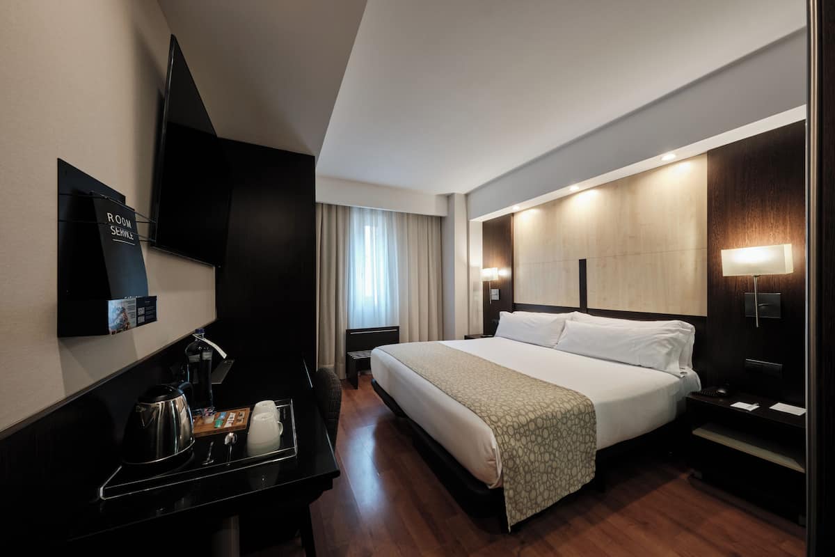 Quarto de hotel moderno com uma cama grande, uma escrivaninha com chaleira, televisão de tela plana e janela com cortinas.