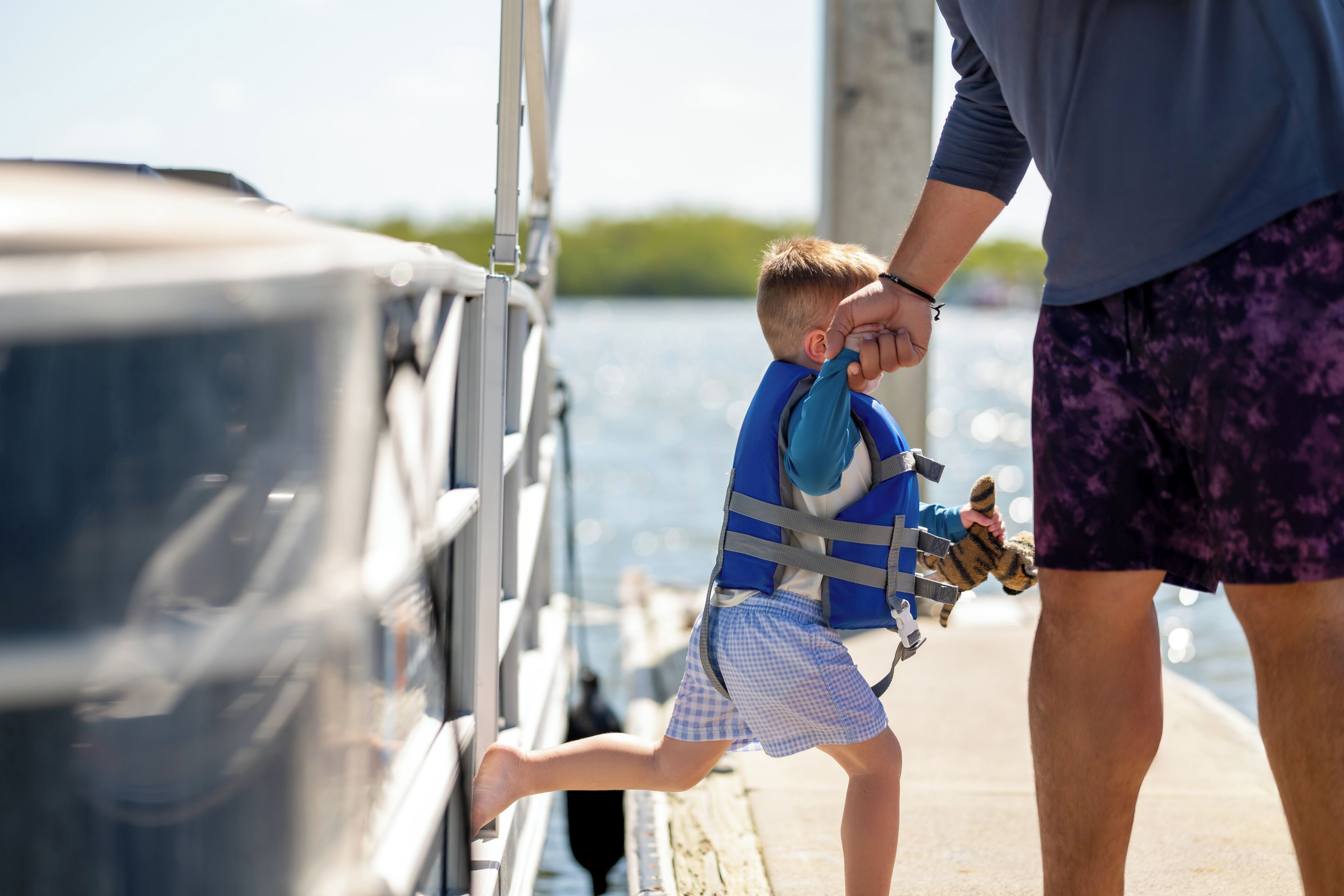A child in a life jacket on a dock, a boat, and a person standing nearby.