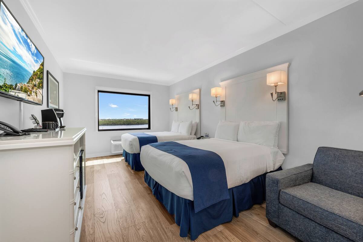 A hotel room with two beds, a desk with a telephone, a grey sofa, and a large window with a view of the sea.
