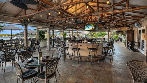 An outdoor bar area with bamboo ceiling beams, multiple tables and chairs, and a TV screen. - Bayfront Inn Fifth Avenue (Naples)