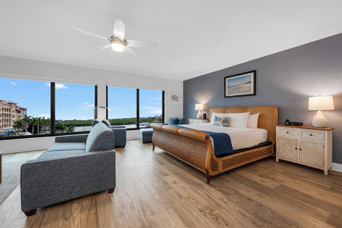 A modern bedroom with a large bed, a sofa, a nightstand, and a view of the city.