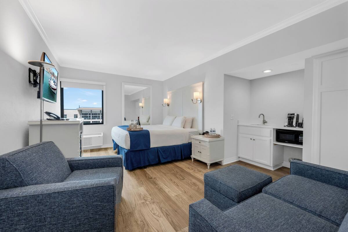 A modern hotel room with a bed, two seating areas, a kitchenette, and a large window offering a view.