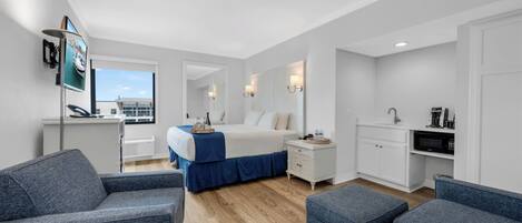 A modern hotel room with a bed, two seating areas, a kitchenette, and a large window offering a view.