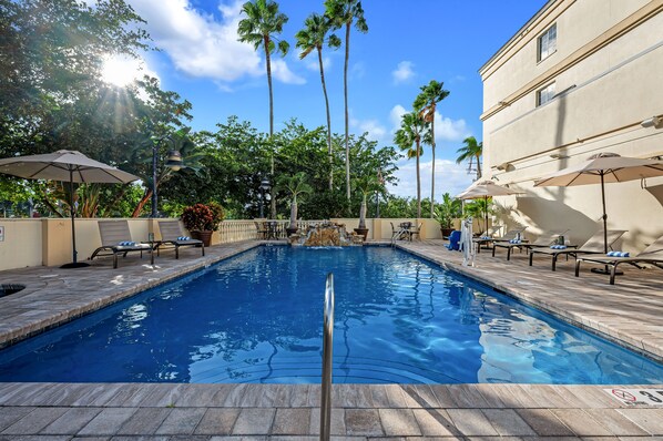 A swimming pool with lounge chairs and umbrellas, surrounded by palm trees and a building.