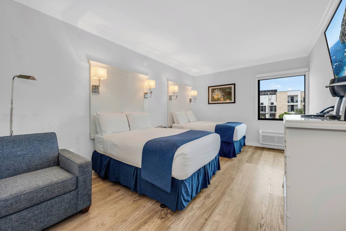 A hotel room with two beds, a sofa, a desk, and a large window with a view of buildings.