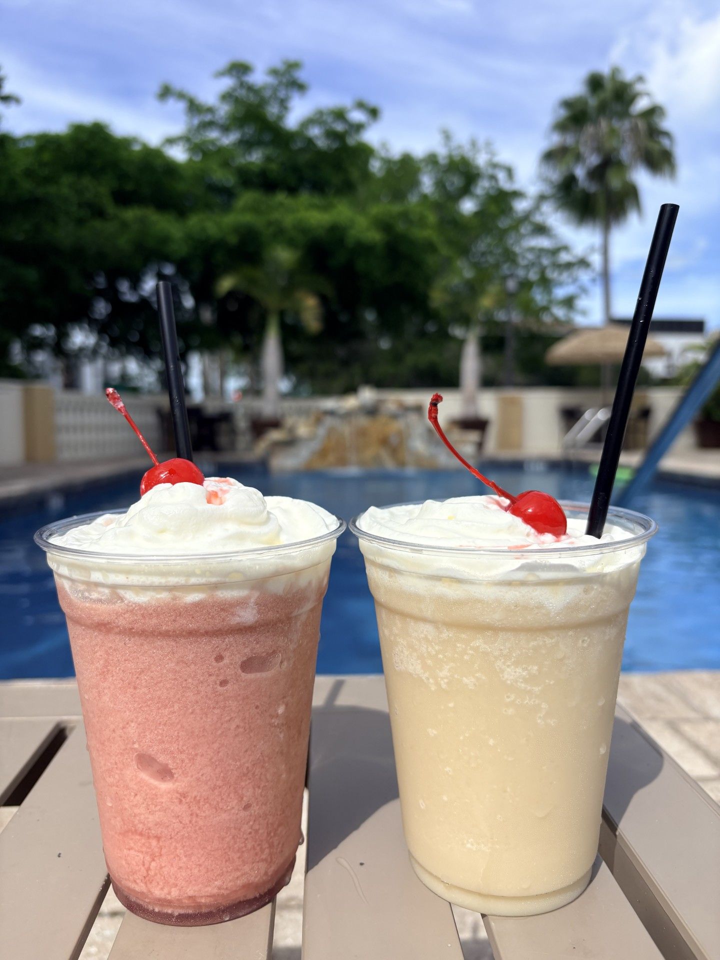 Two beverages with whipped cream and cherries on a table by a poolside.