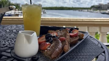 Un plato de waffles con fruta y una bebida sobre una mesa, con vista al agua y al cielo.