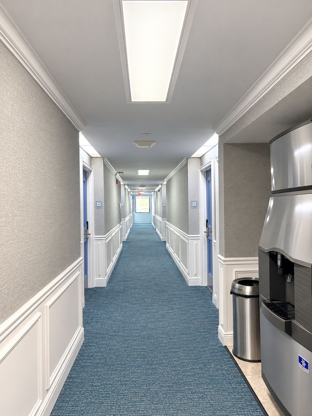 A hotel corridor with a blue carpet, white walls, and a stainless steel refrigerator.
