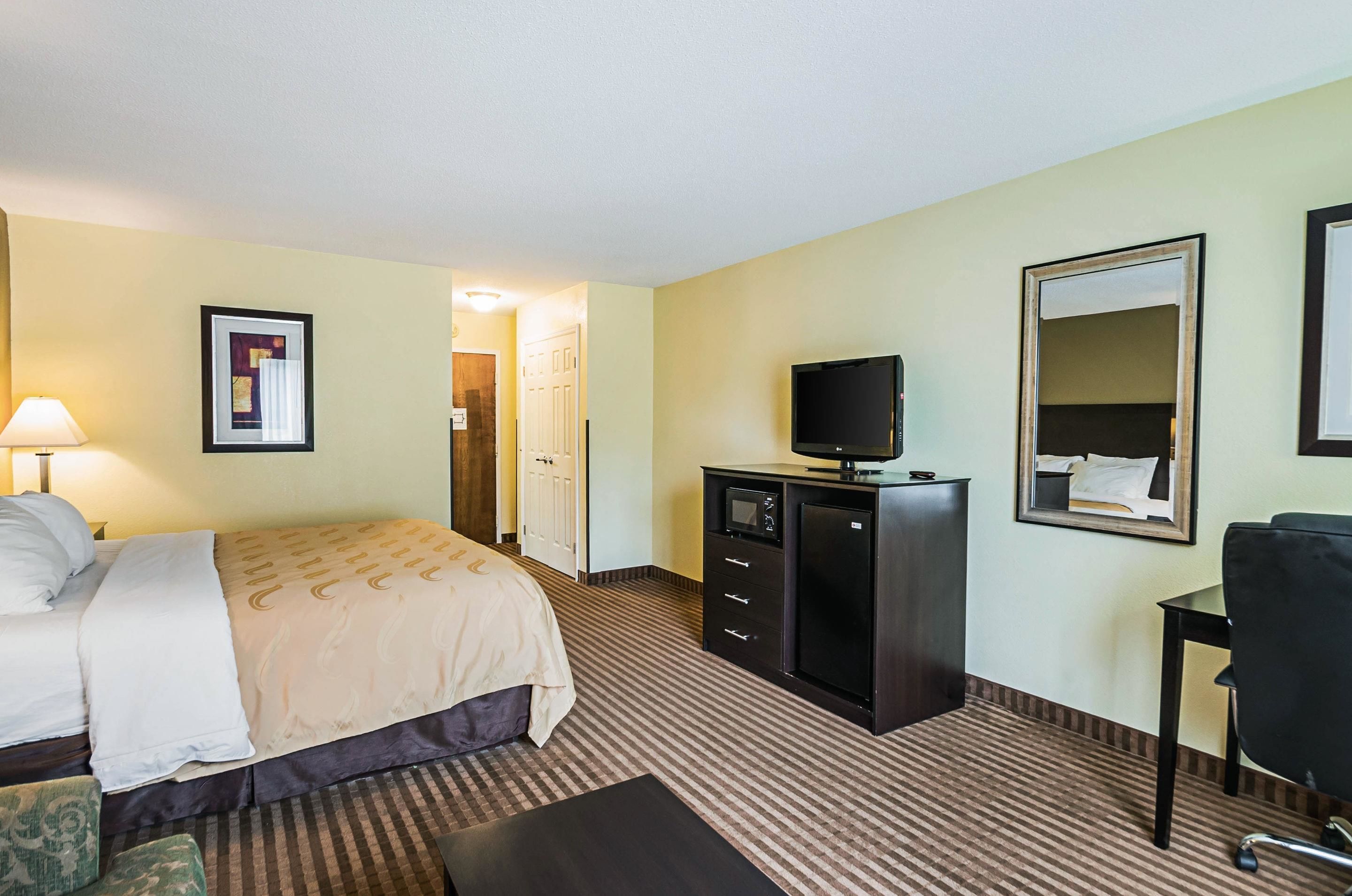 A hotel room with a bed, a desk, a television, a mirror, and a door leading to another room.