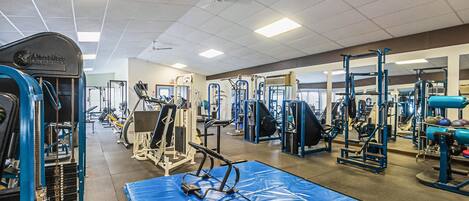 Un gimnasio con diversos equipos de ejercicio, entre ellos máquinas de pesas y pesas libres, y una colchoneta azul en el suelo.