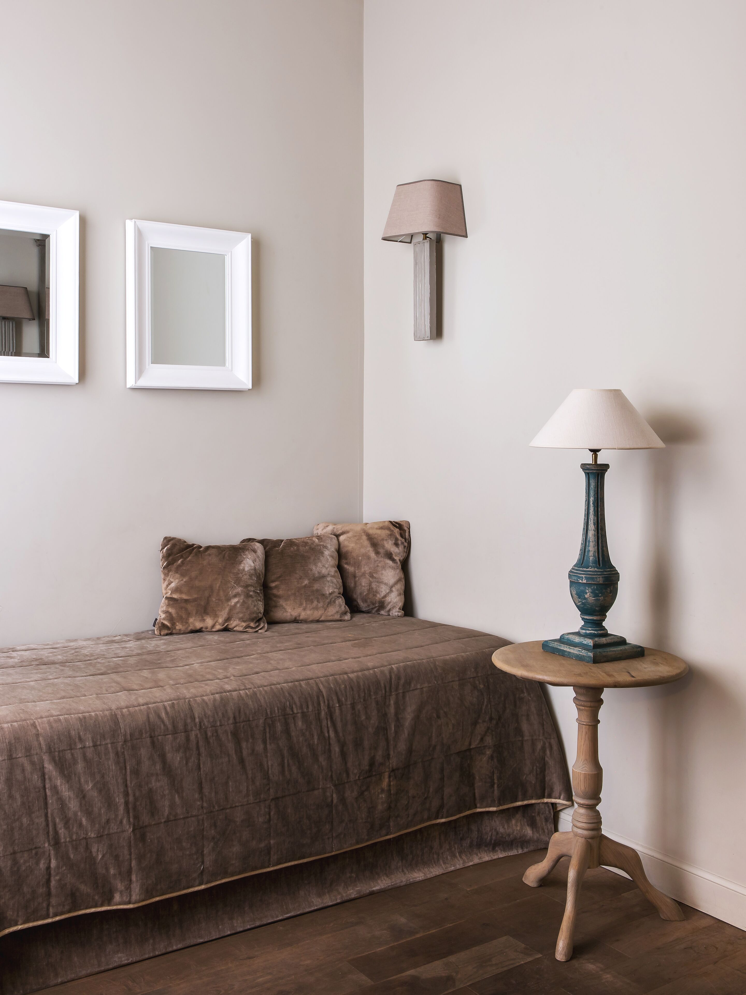 Une chambre à coucher avec un lit, deux tableaux encadrés, une lampe fixée au mur, une table ronde avec une lampe et une tête de lit marron recouverte de tissu.