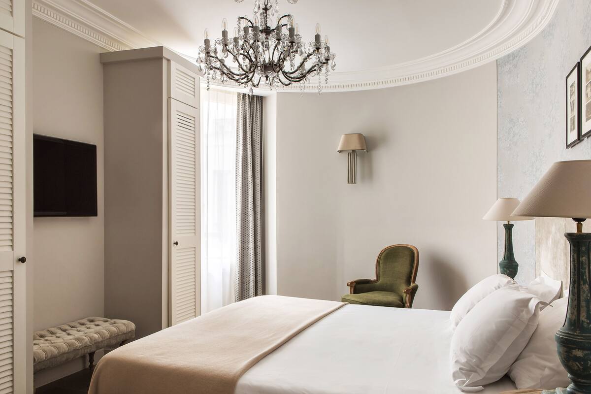 A bedroom with a bed, a bench, a chair, a chandelier, and a lamp.