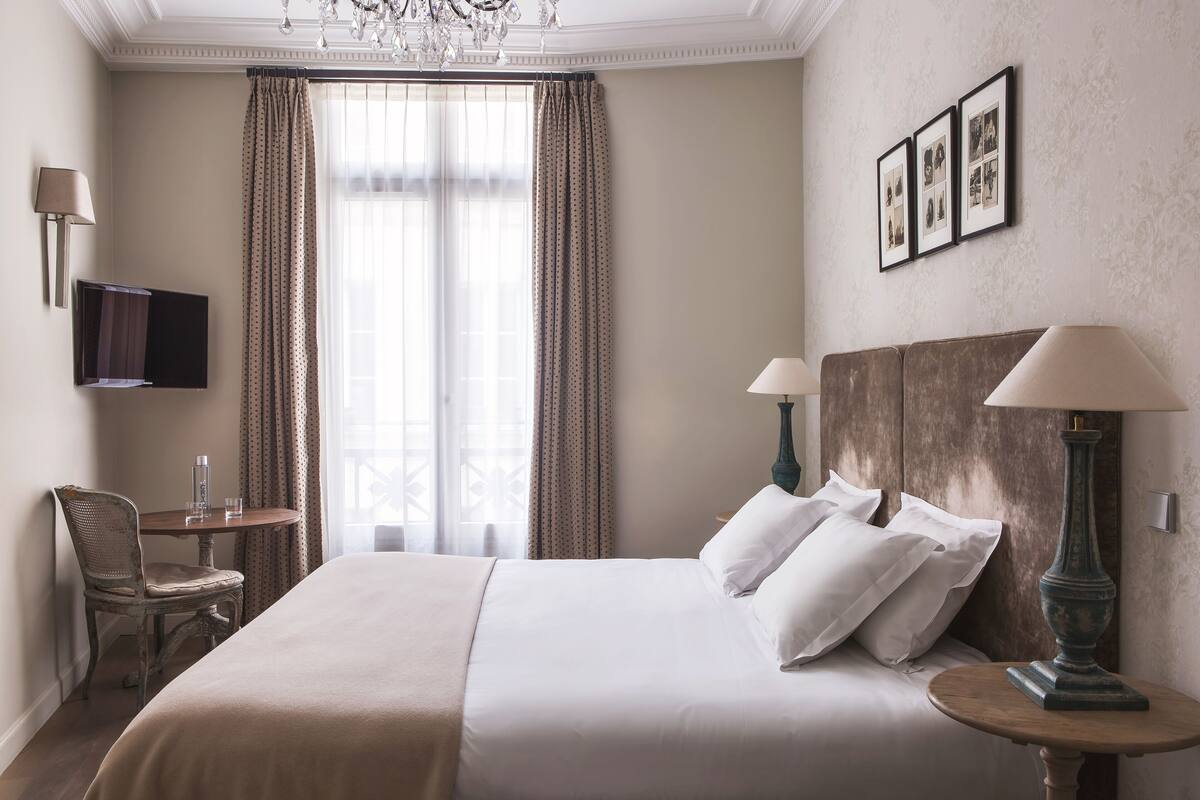 A hotel room with a large bed, two bedside lamps, a small table with a chair, a TV mounted on the wall, and a window with curtains.
