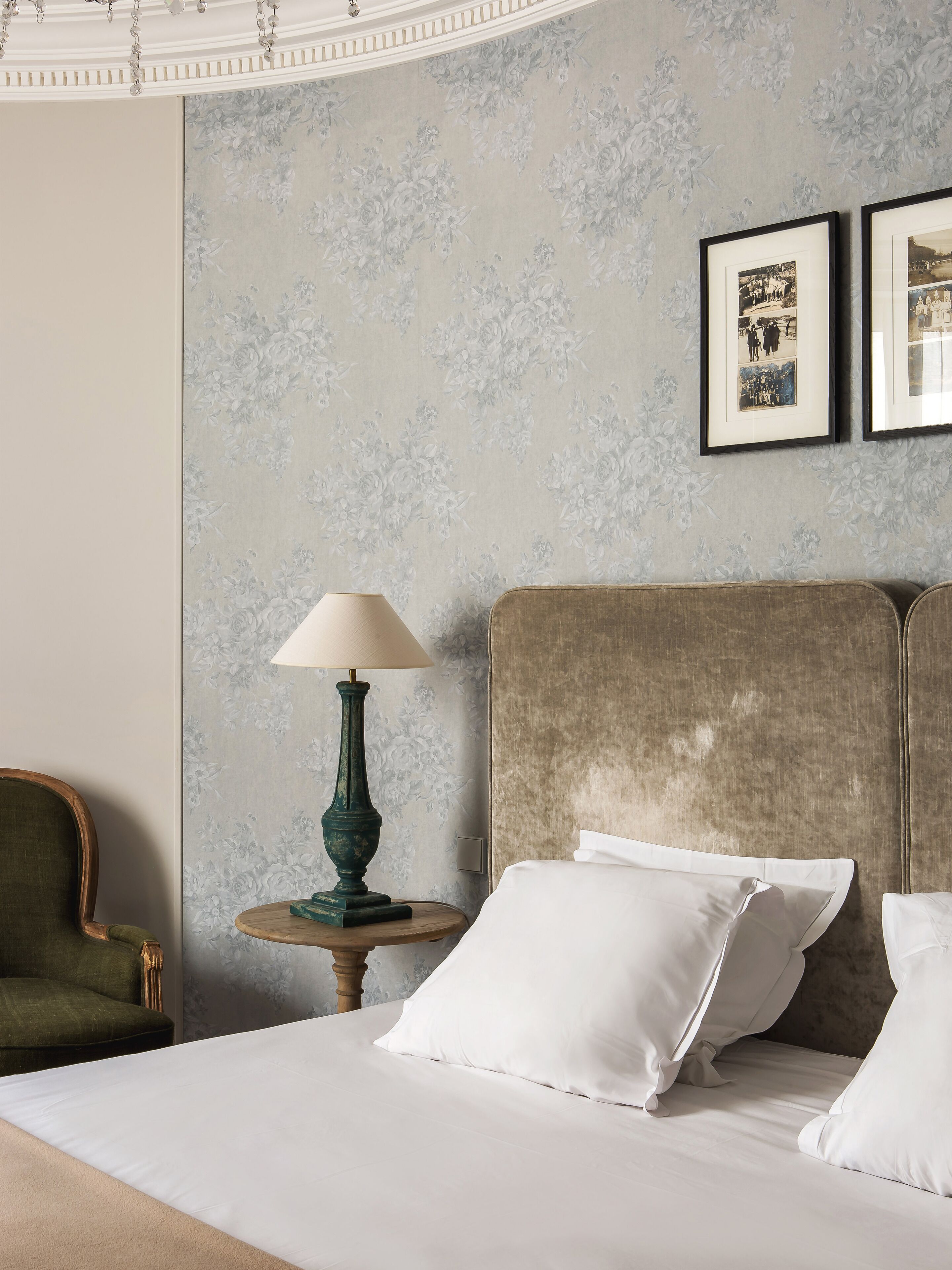 A neatly made bed with white linens, a green upholstered armchair, a round wooden side table with a green lamp, and a textured wall with floral wallpaper.