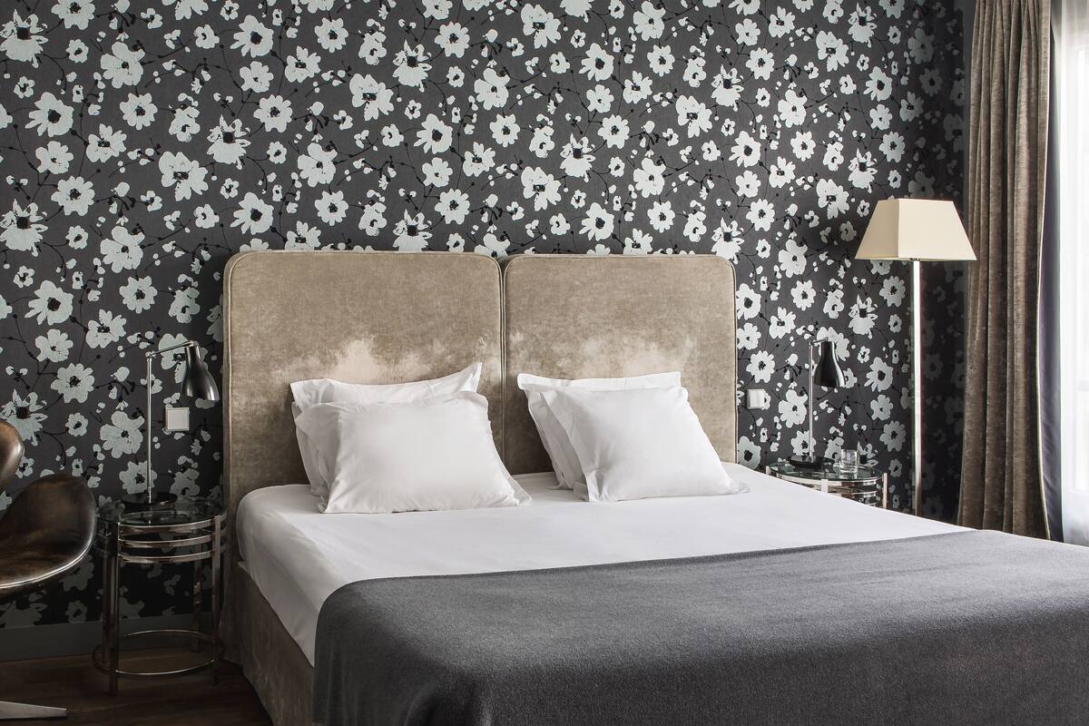 A hotel room with a bed, two pillows, a bedside lamp, a chair, and a wall with a floral pattern.