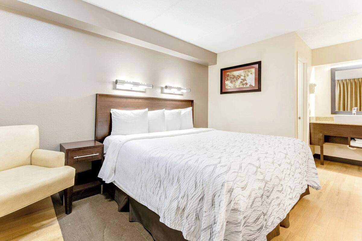 A hotel room with a bed, bedside table, chair, and a bathroom with a mirror and sink.
