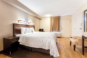 A hotel room with a large bed, a bedside table, a chair, a desk, a window with curtains, and an air conditioning unit. - Red Roof Inn PLUS+ Mt Pleasant - Patriots Point (Mount Pleasant)