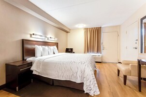 A hotel room with a large bed, a bedside table, a chair, a desk, a window with curtains, and an air conditioning unit.